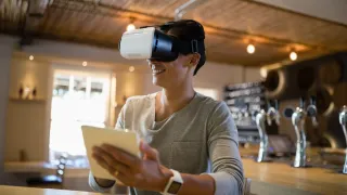 A photo of a person wearing a virtual reality headset is holding a tablet.