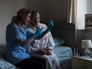 A nurse and senior person are sitting side by side on a bed, looking at a test sample that the nurse is holding
