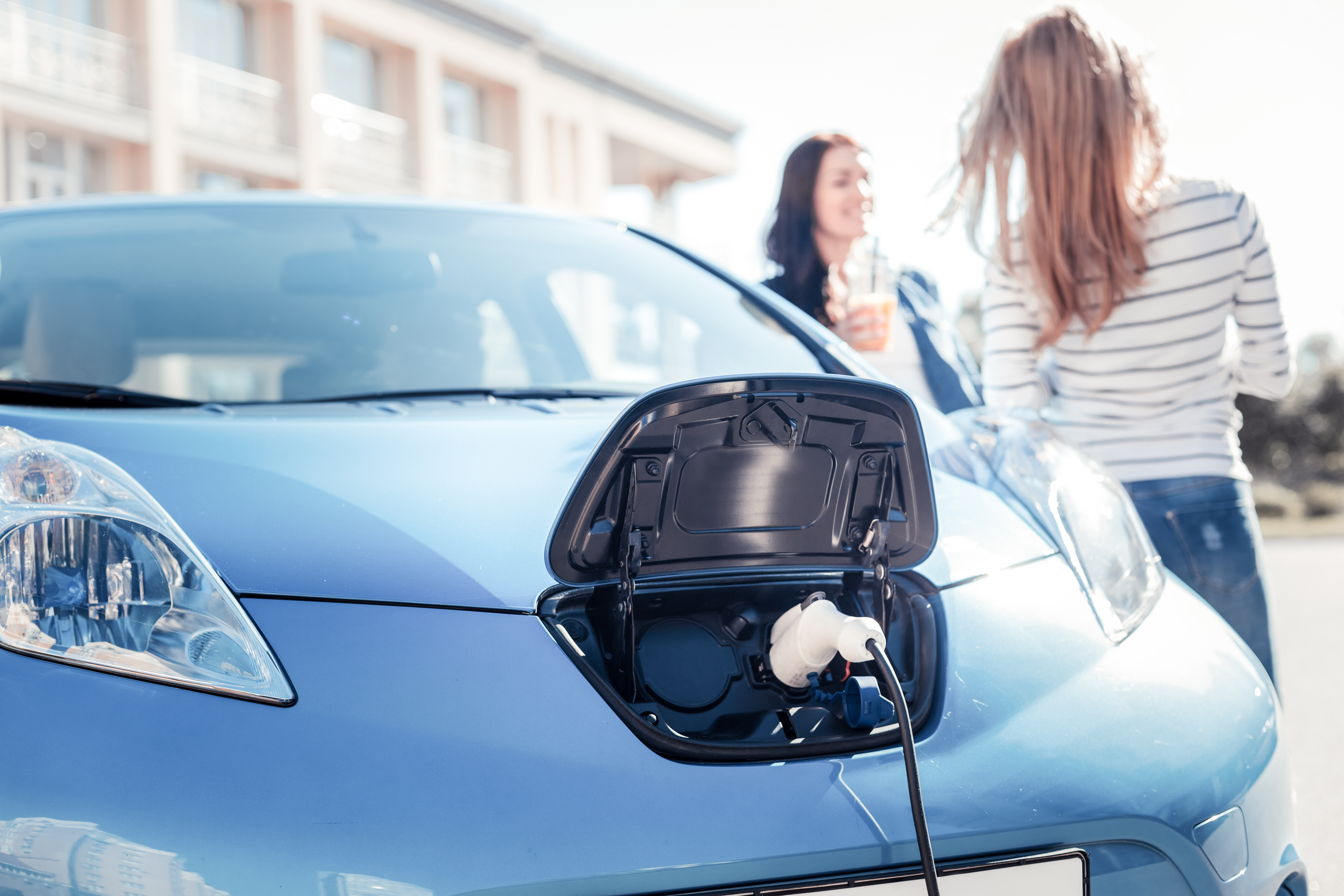 A photo of an electric car being charged, two women are talking in the background.