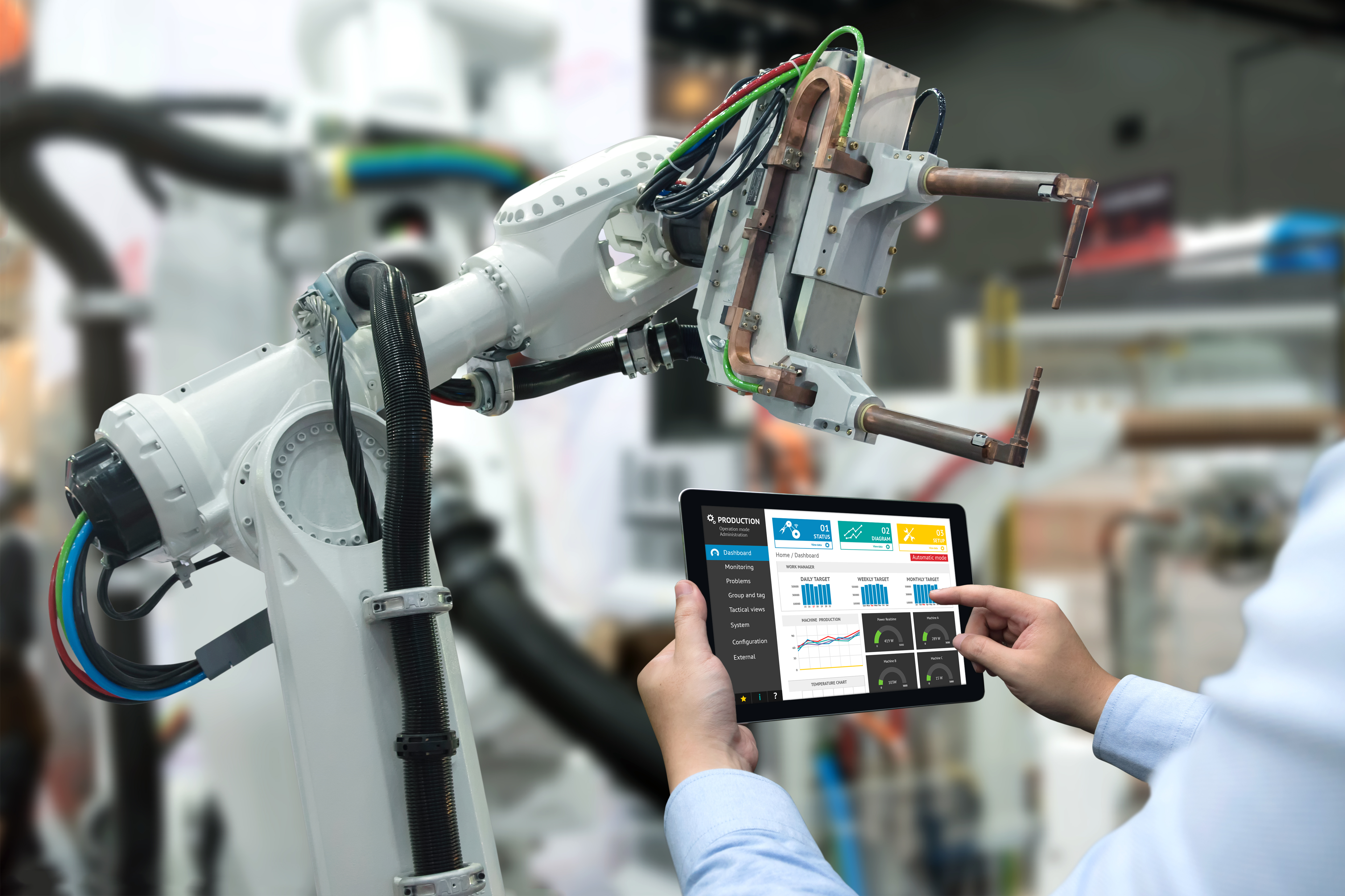 A photo of a robot in a factory setting. A person is holding up a tablet next to in, looking at graphs.