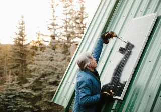 A manw ith grey hair is screwing a solar panel to a house, forest trees in the background