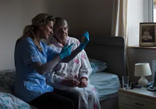 A nurse and senior person are sitting side by side on a bed, looking at a test sample that the nurse is holding