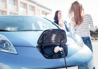 A photo of an electric car being charged, two women are talking in the background.