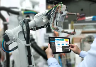 A photo of a robot in a factory setting. A person is holding up a tablet next to in, looking at graphs.