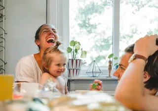 A family of two adults and a small child are sitting in the kitchen, laughing