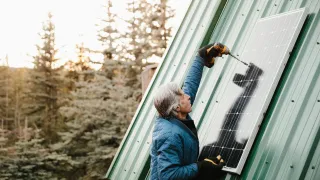 A manw ith grey hair is screwing a solar panel to a house, forest trees in the background