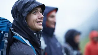 People in weather resistant clothes are standing in the rain and the man in the front is smiling