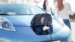 A photo of an electric car being charged, two women are talking in the background.