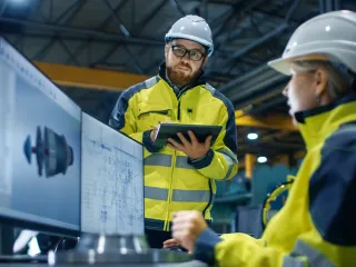 Two persons are looking at schematics on a computer in an industrial setting.