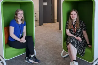 Emma Mulhern and Laura Kojo sitting on green chairs at the office, smiling at the camera.
